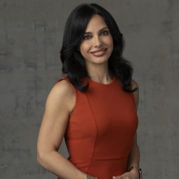Woman with long dark hair wearing a sleeveless orange dress, silver bracelet, and watch, standing against a gray textured background, smiling at the camera.
