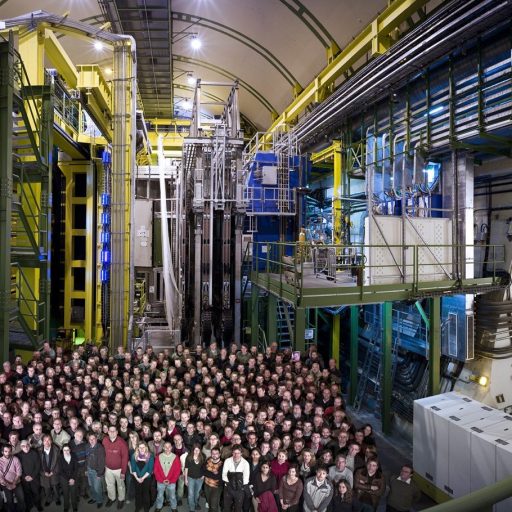 A large group of people stands together inside a spacious, industrial facility—likely the LHC—surrounded by tall machinery, pipes, and metal structures, celebrating the best 2025 discovery in particle physics.