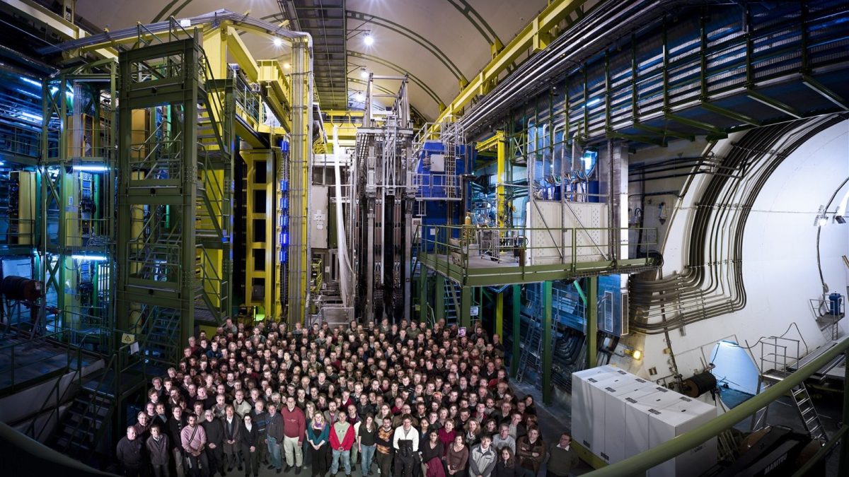 A large group of people stands together inside a spacious, industrial facility—likely the LHC—surrounded by tall machinery, pipes, and metal structures, celebrating the best 2025 discovery in particle physics.