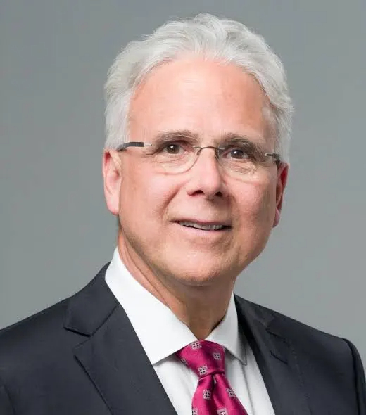 An older man with white hair and glasses wearing a dark suit, white shirt, and pink patterned tie, posing against a plain gray background.