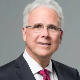 An older man with white hair and glasses wearing a dark suit, white shirt, and pink patterned tie, posing against a plain gray background.
