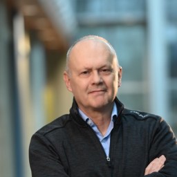 A middle-aged man with short grey hair stands indoors with his arms crossed, wearing a dark zip-up jacket over a light blue shirt. The background is blurred.