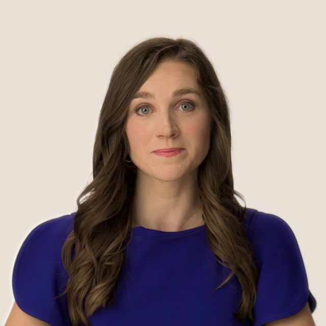 A woman with long brown hair, wearing a blue top, looks directly at the camera with a neutral expression against a plain light background.