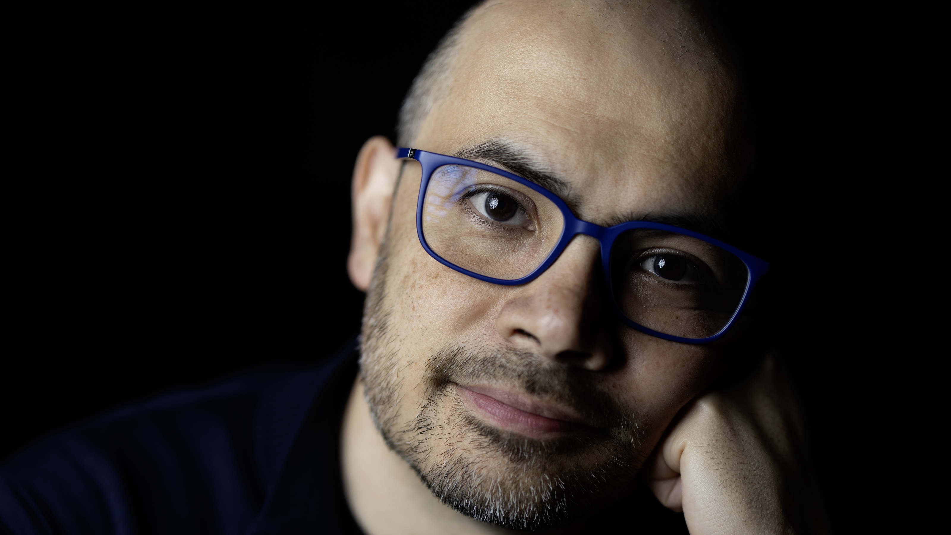 A person with a shaved head, beard, and blue-framed glasses—reminiscent of Demis Hassabis—looks at the camera with their hand resting on their face against a dark background.