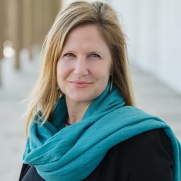 A woman with long blonde hair wearing a teal scarf and black top stands outdoors with a blurred background.
