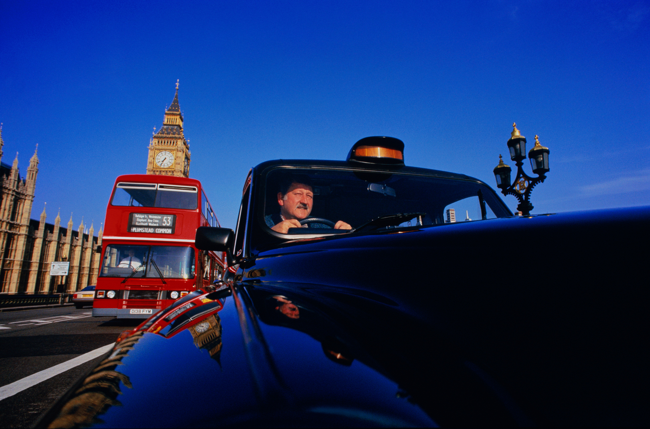 A black cab and a red double-decker bus drive past Big Ben and the Houses of Parliament in London under a clear blue sky.