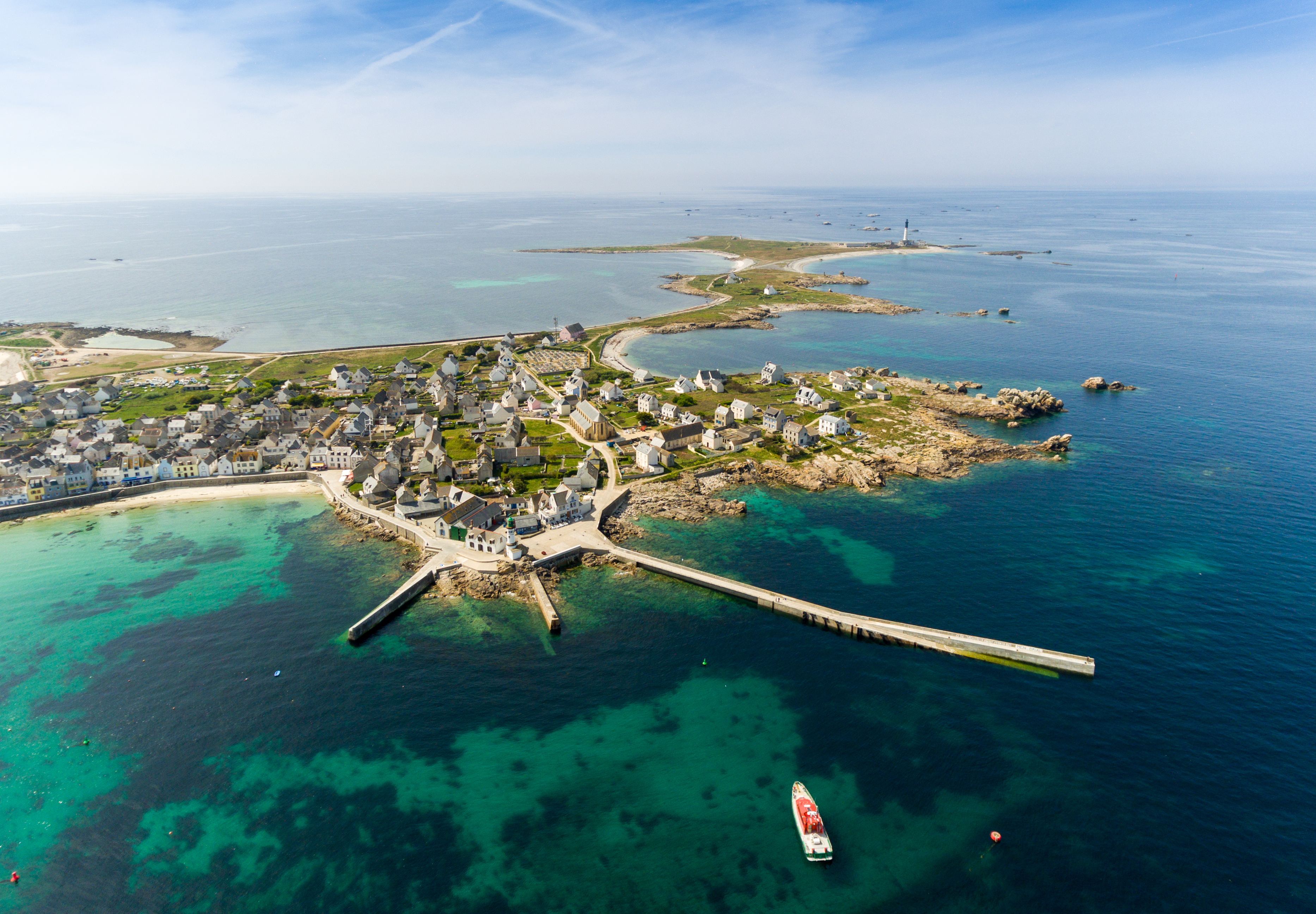 Aerial view of a coastal village with stone houses, a long pier, clear turquoise water, and a red boat near the shore, surrounded by small islands and the open sea.