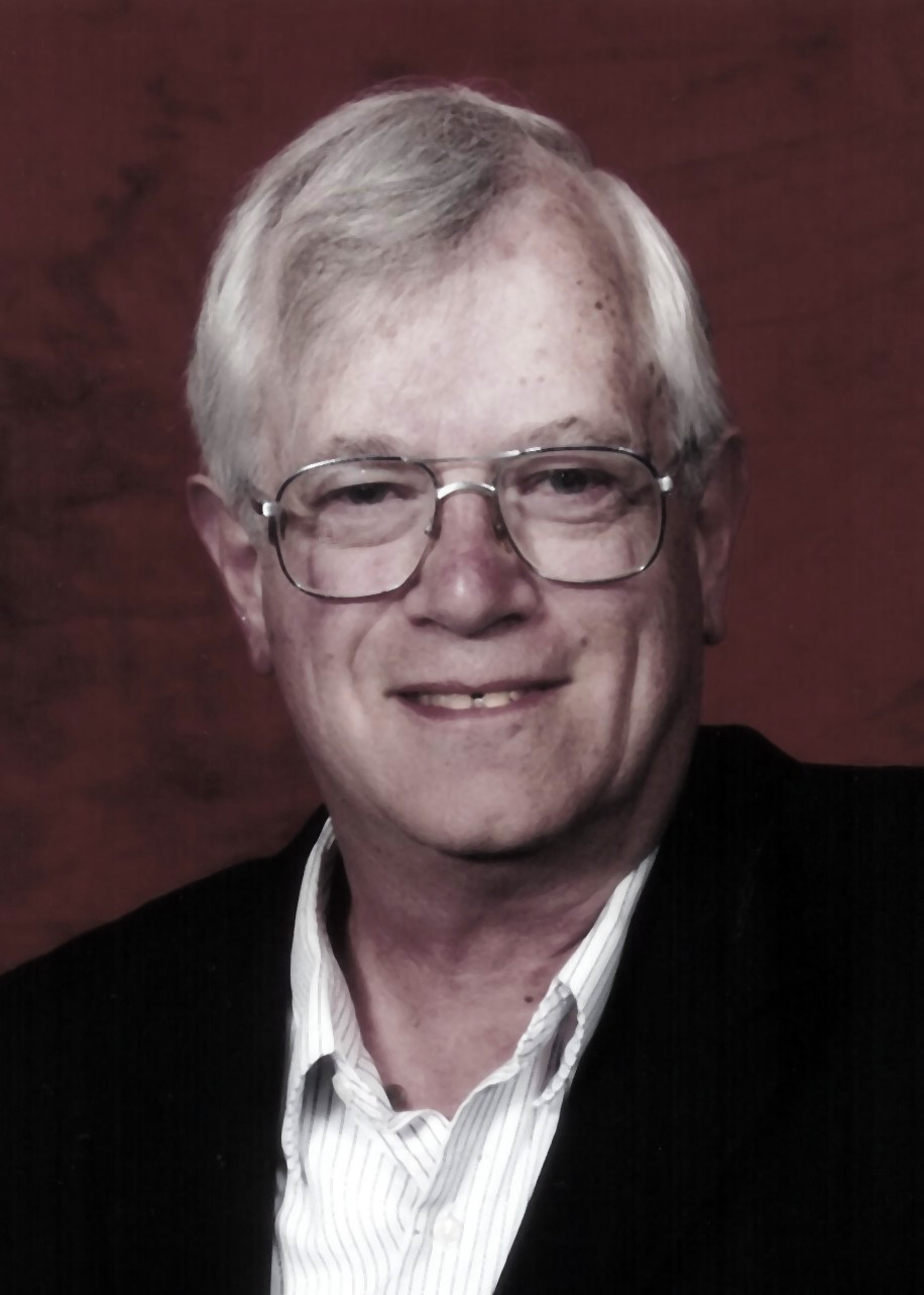 An older man with gray hair and glasses is wearing a dark jacket and a light-collared shirt, posed in front of a reddish-brown background.