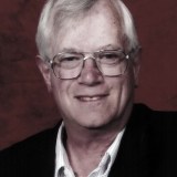 An older man with gray hair and glasses is wearing a dark jacket and a light-collared shirt, posed in front of a reddish-brown background.