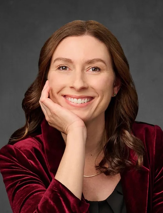 A woman with long brown hair smiles at the camera, resting her chin on her hand. She wears a burgundy jacket and a gold necklace, with a dark gray background behind her.