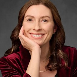 A woman with long brown hair smiles at the camera, resting her chin on her hand. She wears a burgundy jacket and a gold necklace, with a dark gray background behind her.