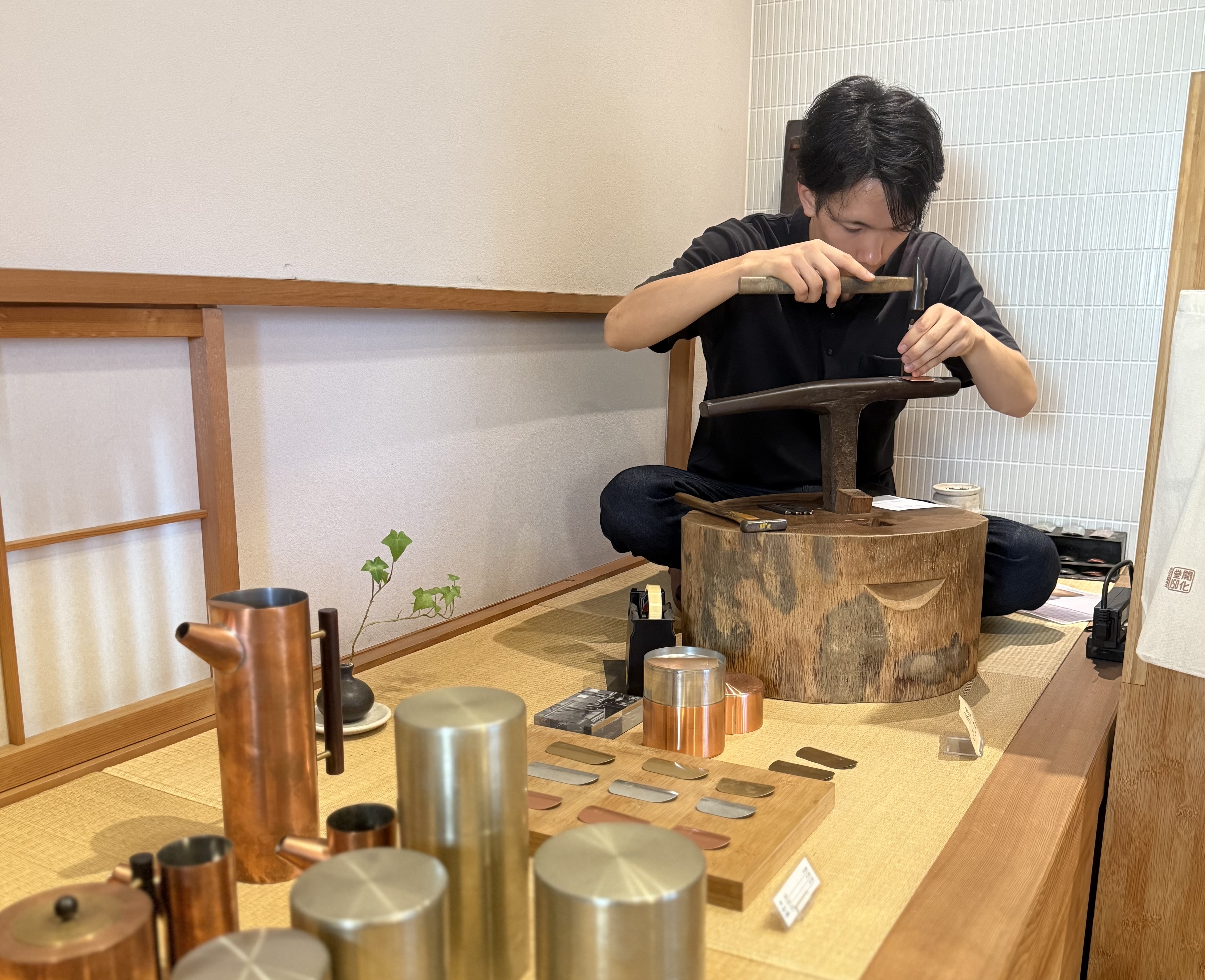 A person sits on a platform, using a hammer and metal tool to work on a dark metal object. Various metal items and tools are displayed on the table in front.