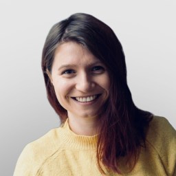A woman with long brown hair wearing a yellow sweater smiles at the camera against a plain light gray background.