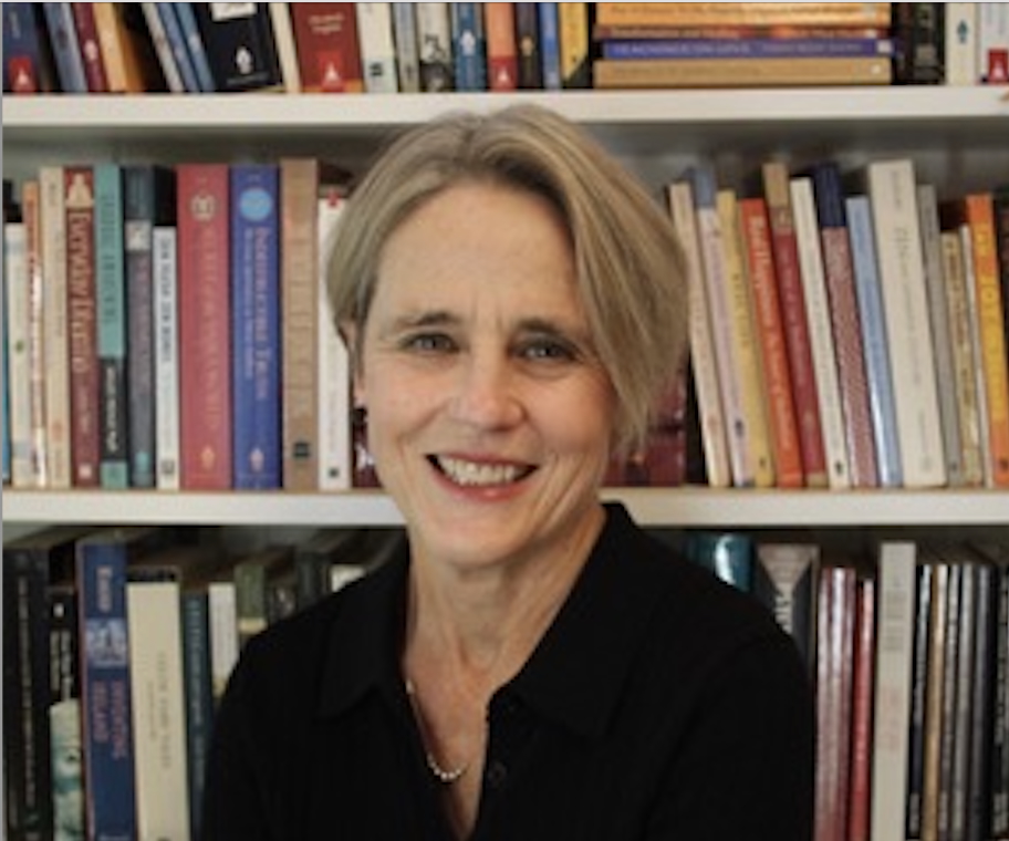A woman with short gray hair and a black top sits and smiles in front of a bookshelf filled with books.