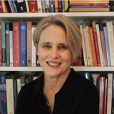A woman with short gray hair and a black top sits and smiles in front of a bookshelf filled with books.