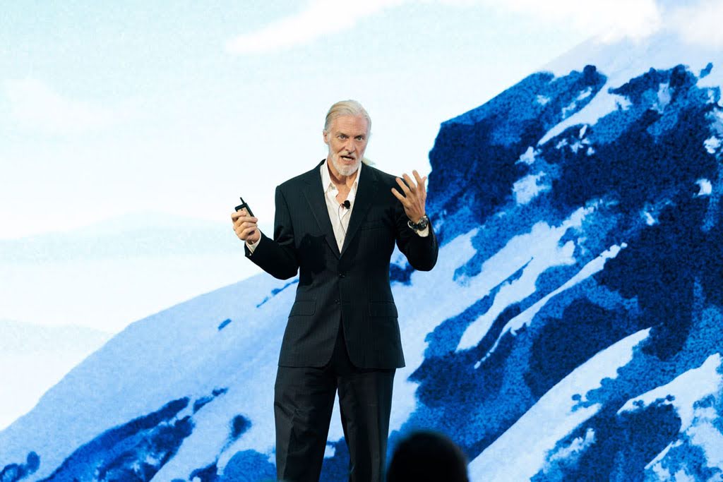 A man in a suit gives a presentation on stage in front of a large image of a snow-covered mountain.