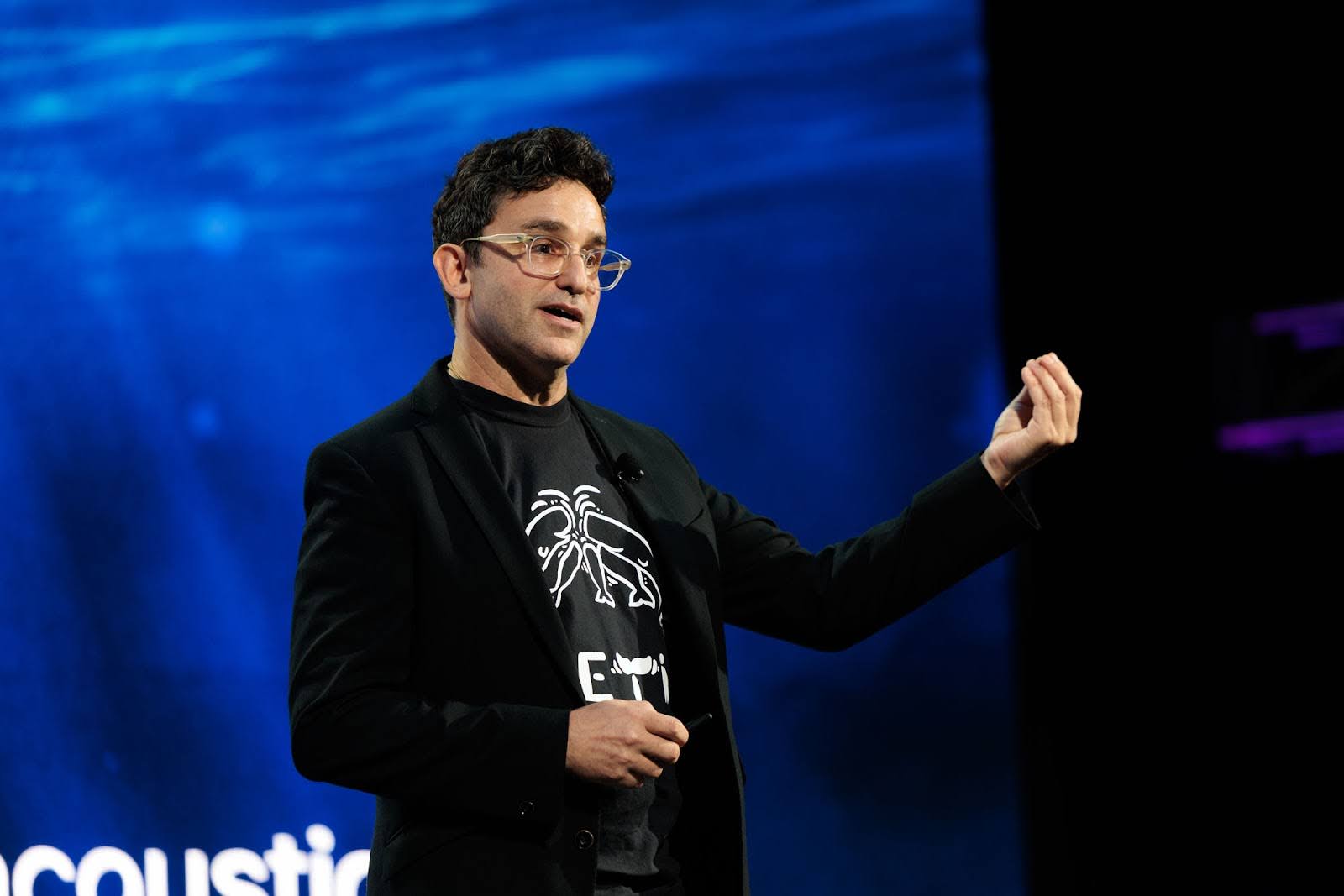 A man wearing glasses and a black blazer gestures while speaking on stage in front of a blue background.