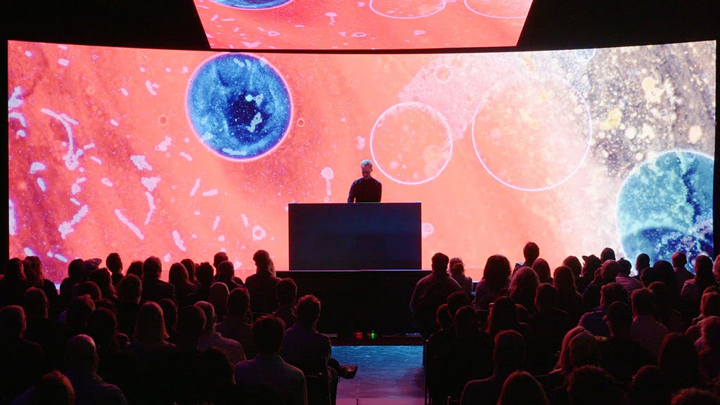 A person stands at a podium on stage before an audience, with large, colorful, abstract visuals displayed on screens behind and above them.