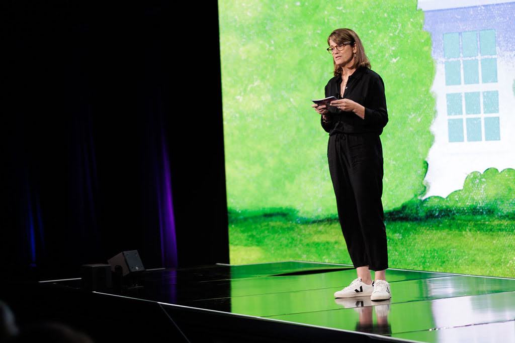A woman in black clothing and white sneakers stands on stage holding note cards, with a green and white background displayed behind her.