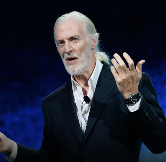 An older man with white hair and beard, wearing a suit and holding a microphone, gestures with one hand while speaking on stage against a dark blue background.