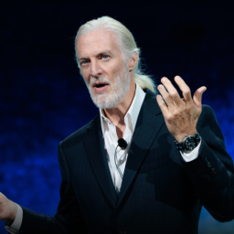 An older man with white hair and beard, wearing a suit and holding a microphone, gestures with one hand while speaking on stage against a dark blue background.