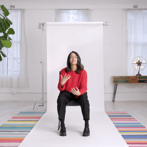 A person in a red sweater sits on a stool in front of a white backdrop in a bright room with plants, a colorful rug, and a table with a lamp.