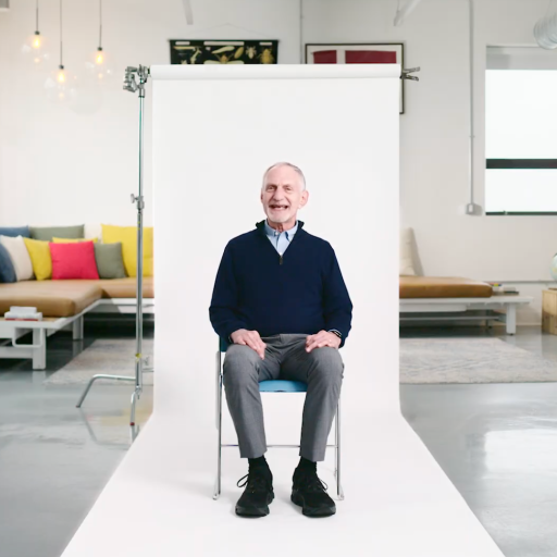 An older man sits on a chair in front of a white backdrop in a modern, brightly lit room with colorful pillows and minimalist decor.