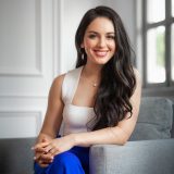 A woman with long dark hair, wearing a white sleeveless top and blue pants, sits on a gray chair and smiles at the camera in a bright room with white walls and large windows.