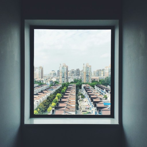 A large square window frames a view of rows of houses and tall apartment buildings under a partly cloudy sky.