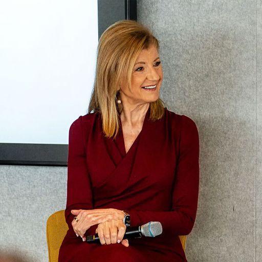 Arianna Huffington, in a maroon dress, sits holding a microphone and smiling in front of a light grey and white background.