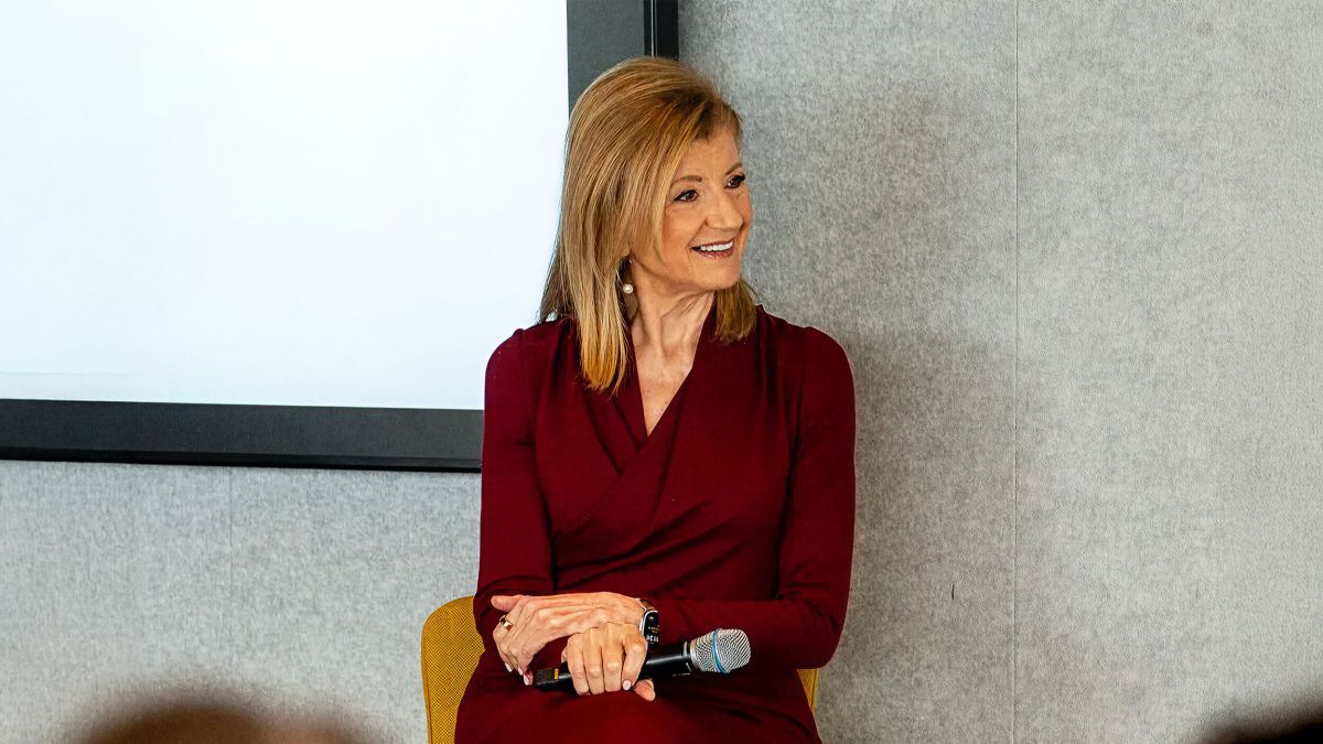 Arianna Huffington, in a maroon dress, sits holding a microphone and smiling in front of a light grey and white background.