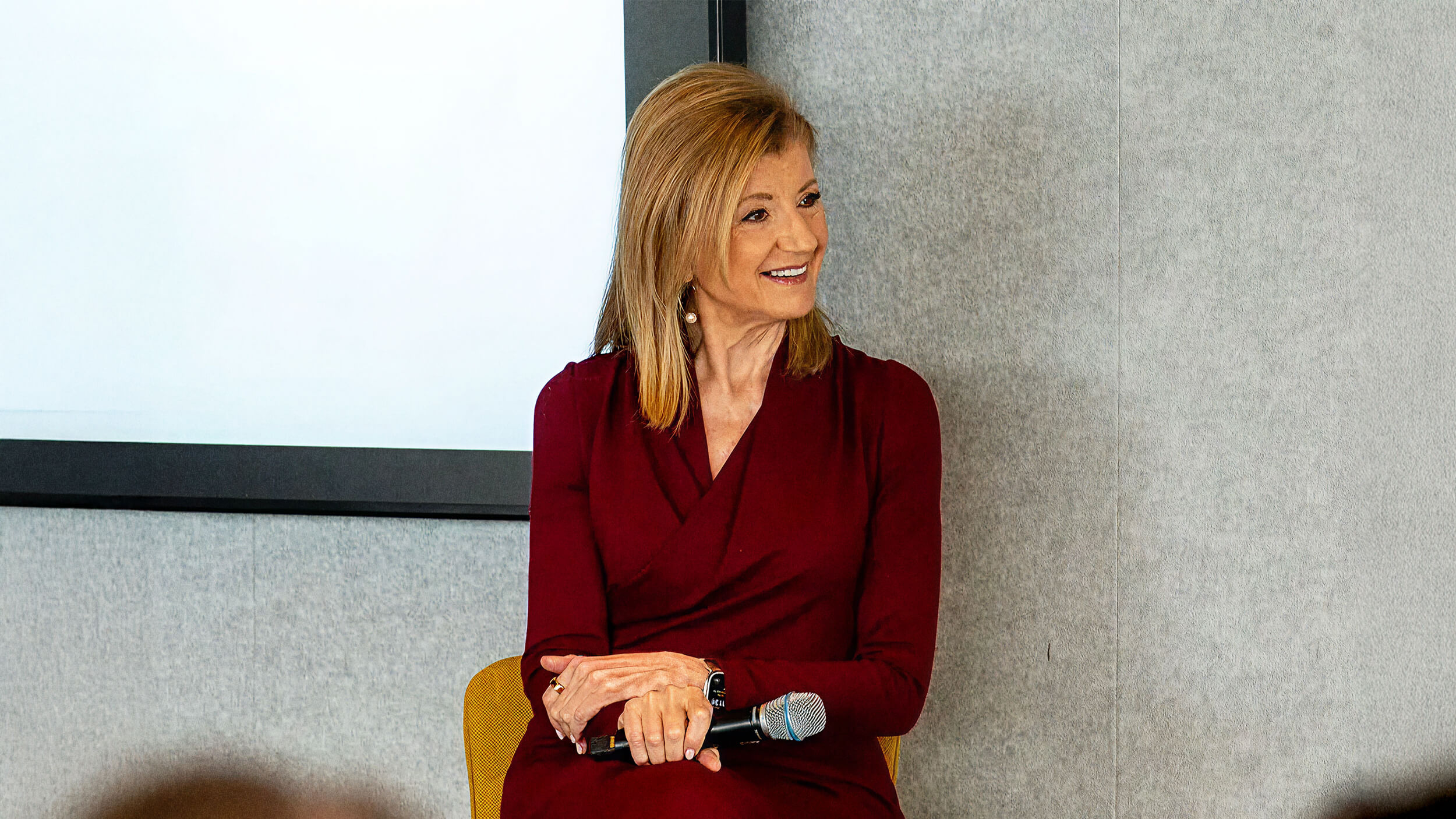 Arianna Huffington, in a maroon dress, sits holding a microphone and smiling in front of a light grey and white background.