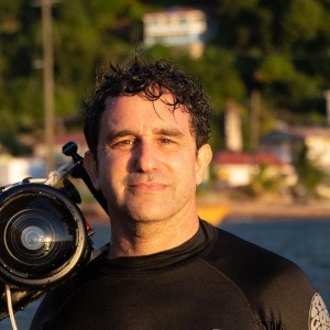 A man in a wetsuit stands outdoors holding a professional camera housing, with water and buildings in the blurred background.