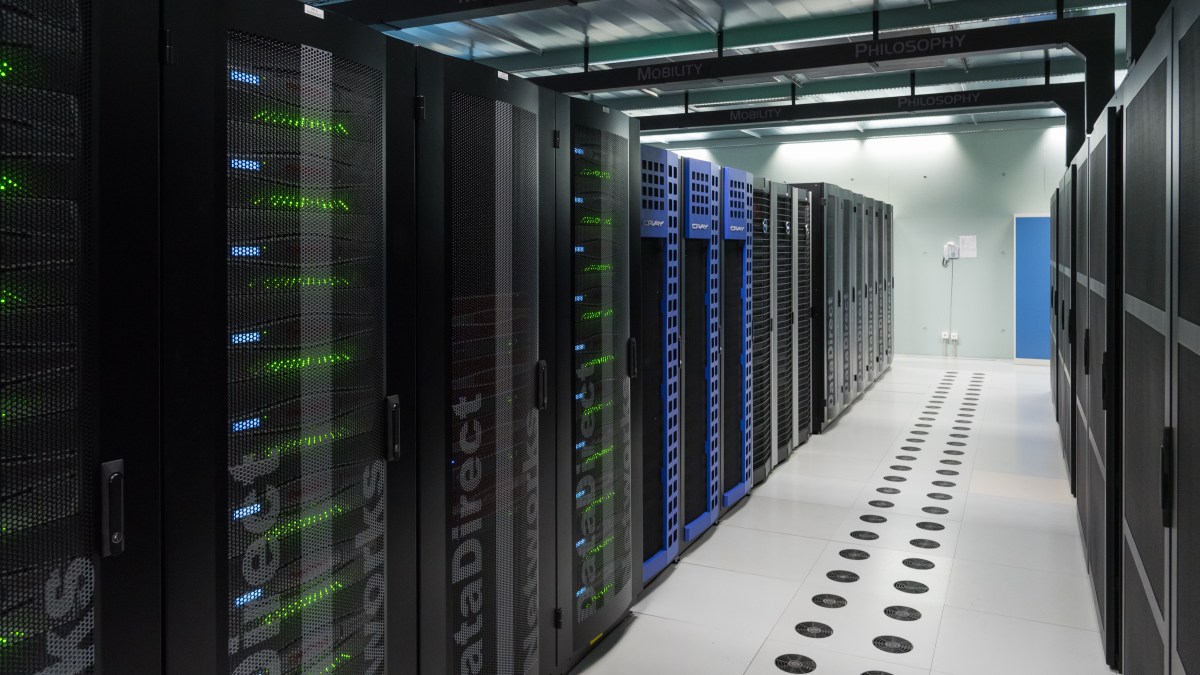 A row of black and blue server racks in a data center, where LLMs power chatbot solutions, with illuminated green lights and a white tile floor with black circular vents.