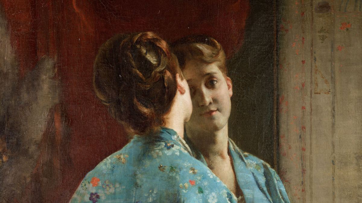 A woman in a blue floral robe looks at her reflection in a mirror, her back partially turned to the viewer as she contemplates her identity.