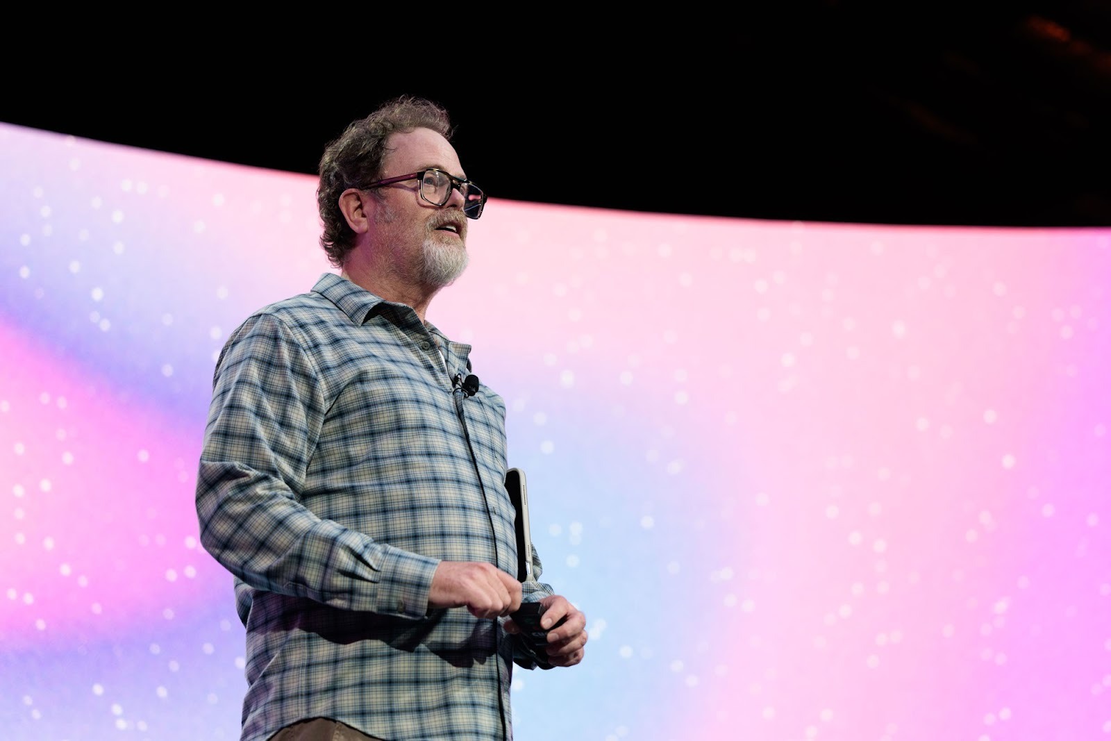 A man in a plaid shirt stands on stage holding a device, speaking in front of a large pink and blue gradient screen with white speckles.