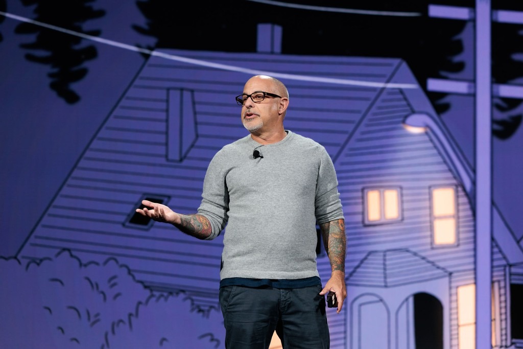 A man with glasses and tattoos, wearing a gray sweater and dark pants, speaks on stage in front of an illustrated house backdrop.