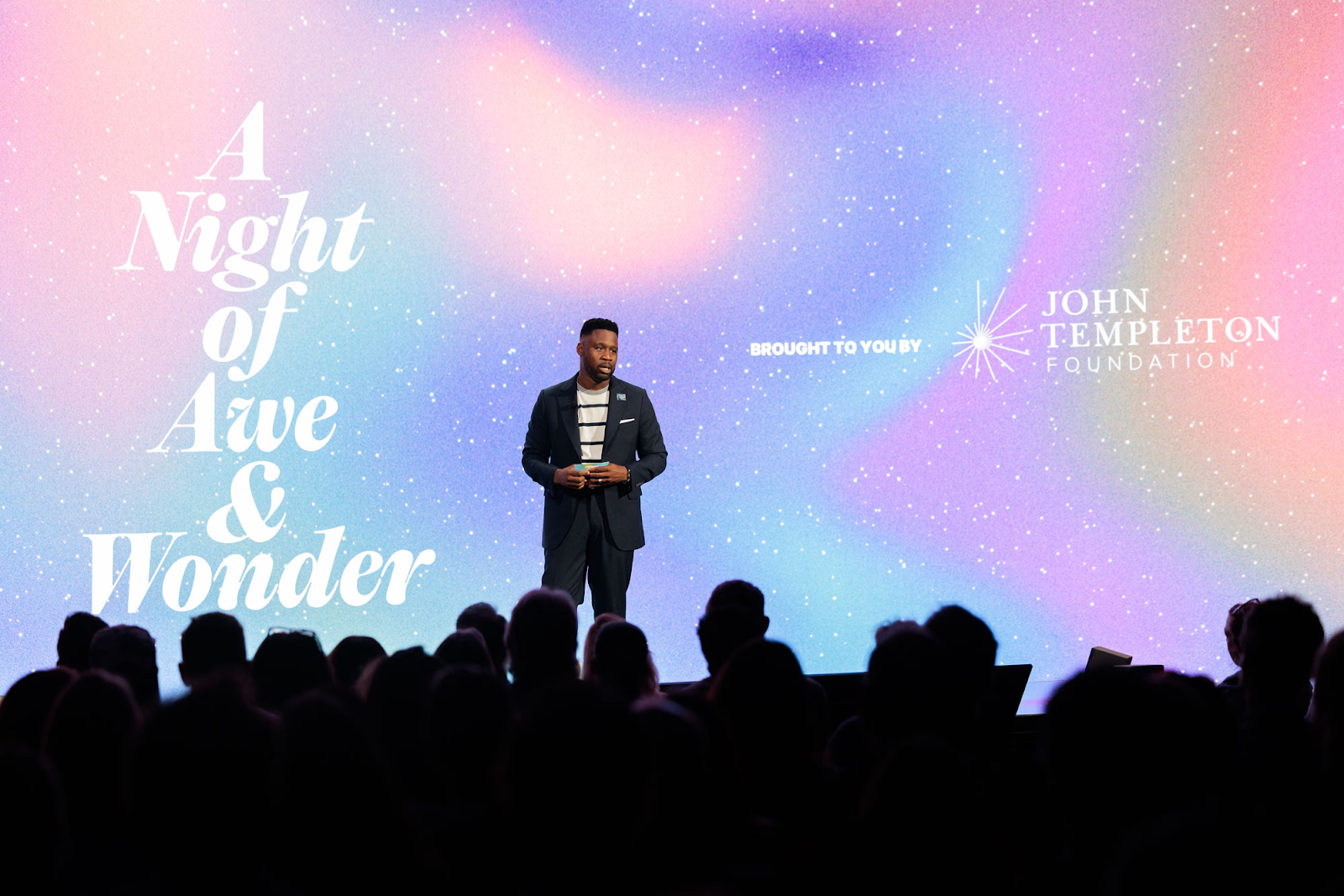 A man stands on stage before an audience, with a backdrop reading "A Night of Awe & Wonder" and the John Templeton Foundation logo.