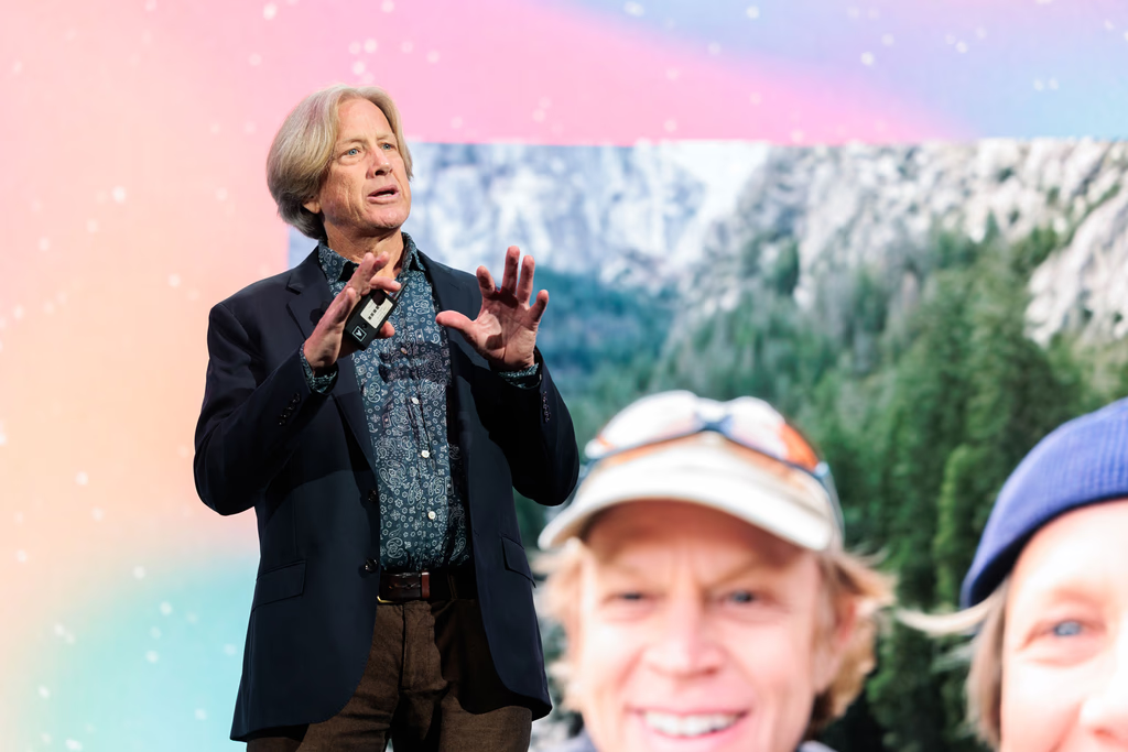 A man in a blazer and patterned shirt speaks on stage in front of a large outdoor-themed background image.