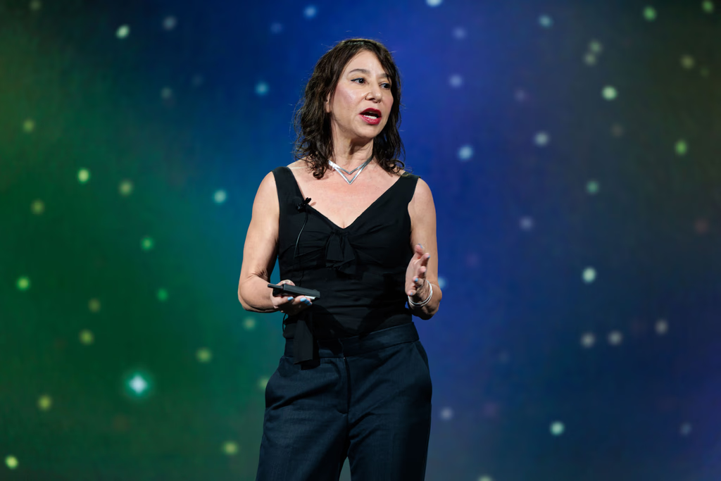 A woman in a black sleeveless top and dark pants speaks on stage against a blue and green starry background, holding a device in one hand.