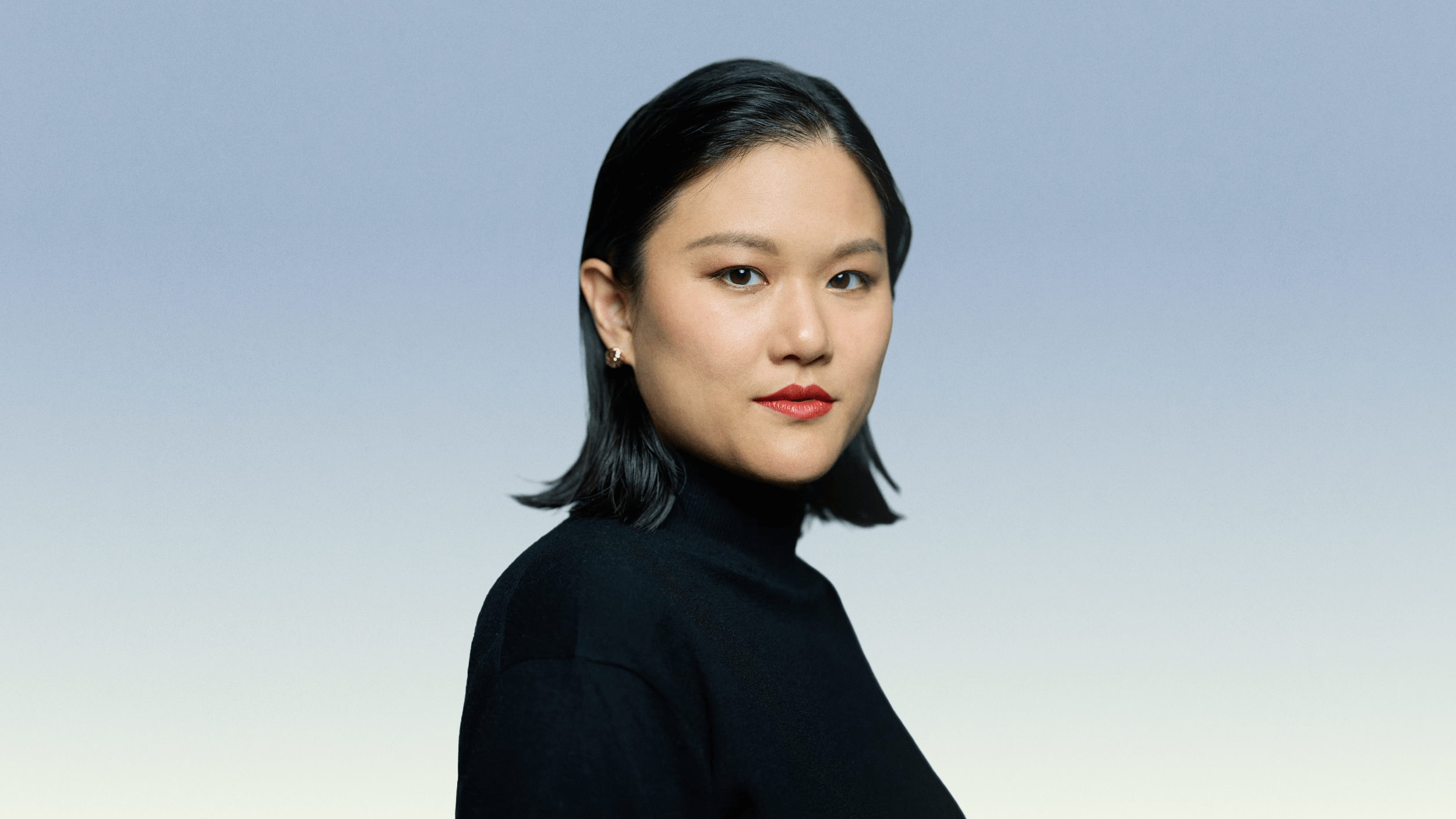 A woman with straight black hair, wearing a black turtleneck, poses against a blue-to-white gradient background.