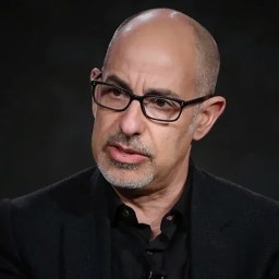 A middle-aged man with glasses and a goatee, wearing a dark suit, speaks against a dark background.