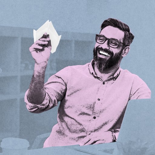 A man with a beard and glasses smiles as he holds a paper airplane in an office setting, savoring life’s simple joys.