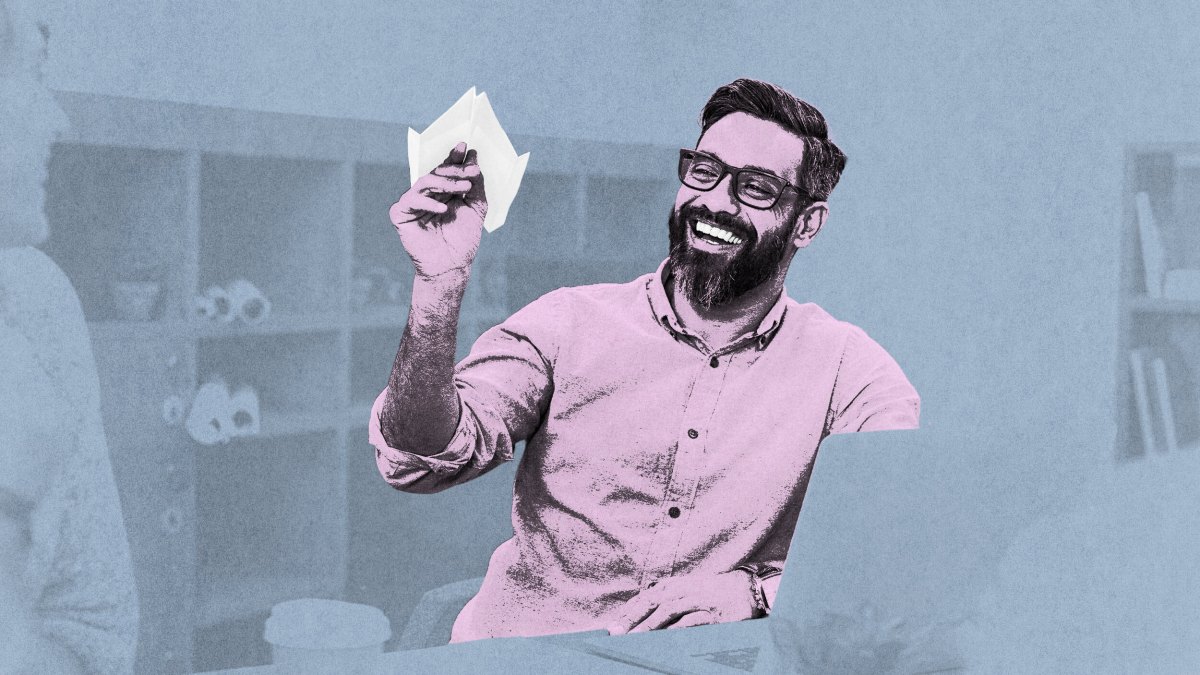 A man with a beard and glasses smiles as he holds a paper airplane in an office setting, savoring life’s simple joys.
