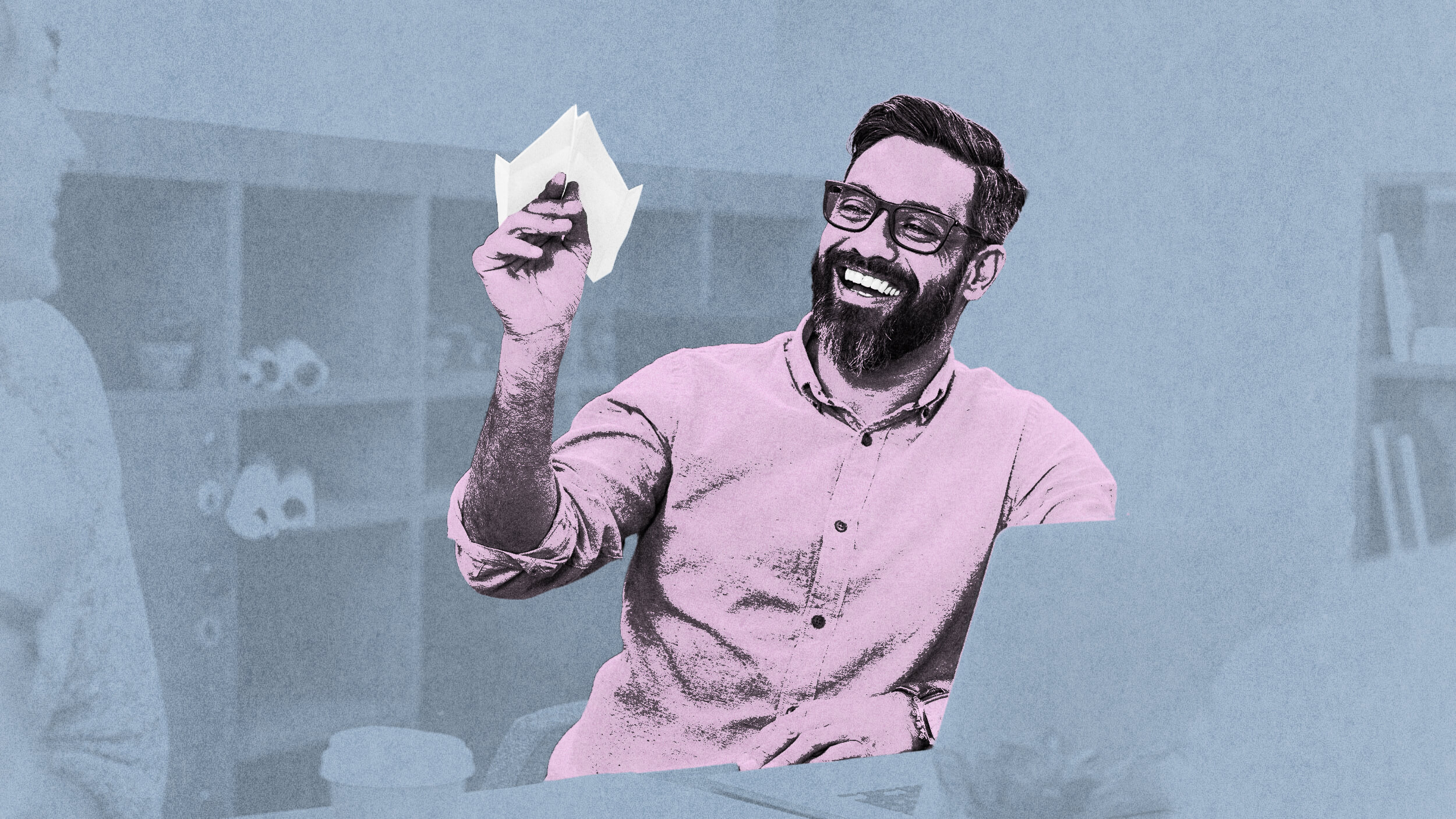 A man with a beard and glasses smiles as he holds a paper airplane in an office setting, savoring life’s simple joys.