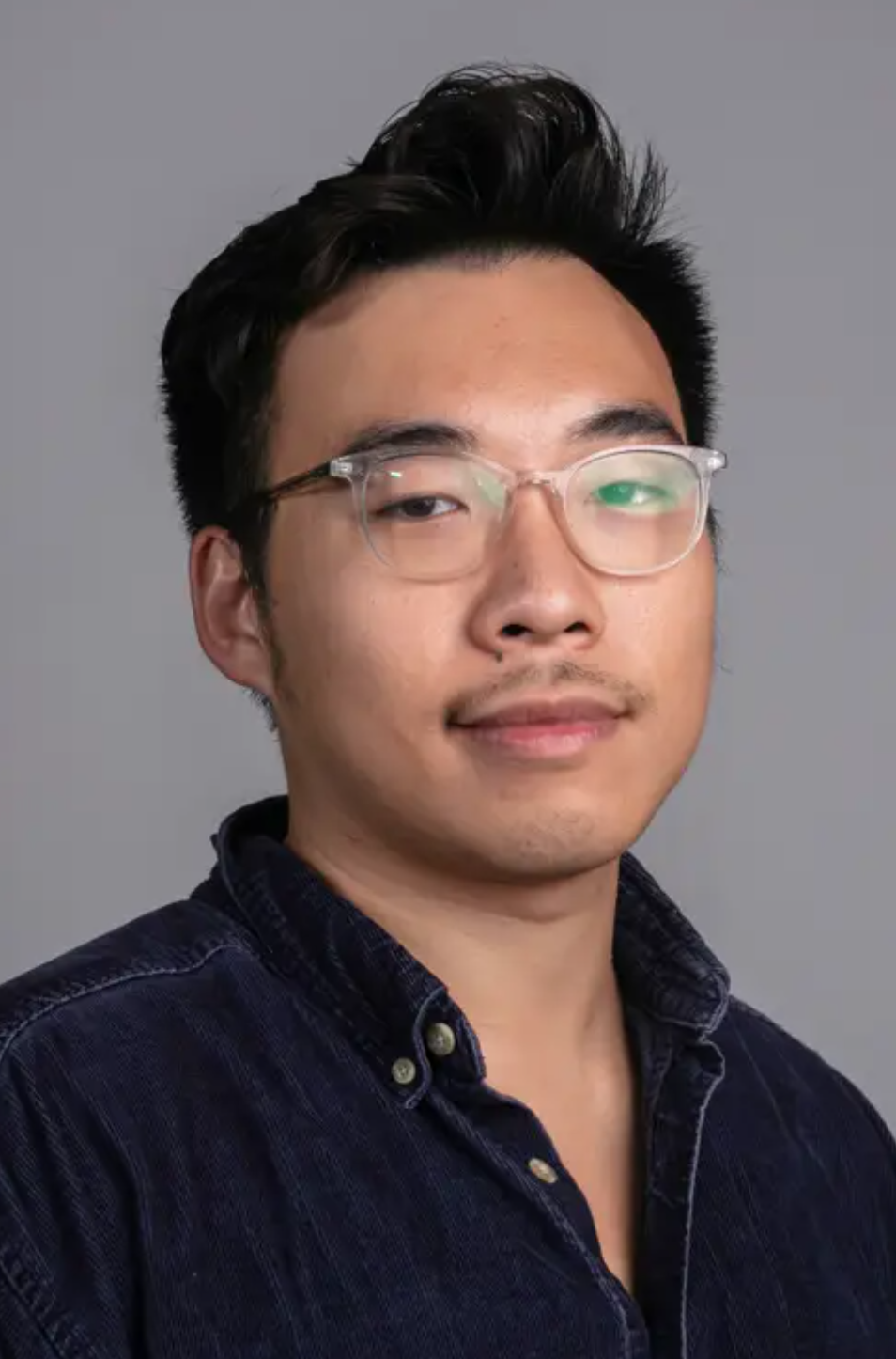 A person with short black hair, glasses, and slight facial hair wearing a dark button-up shirt, posed against a plain grey background.