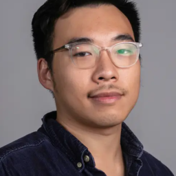 A person with short black hair, glasses, and slight facial hair wearing a dark button-up shirt, posed against a plain grey background.
