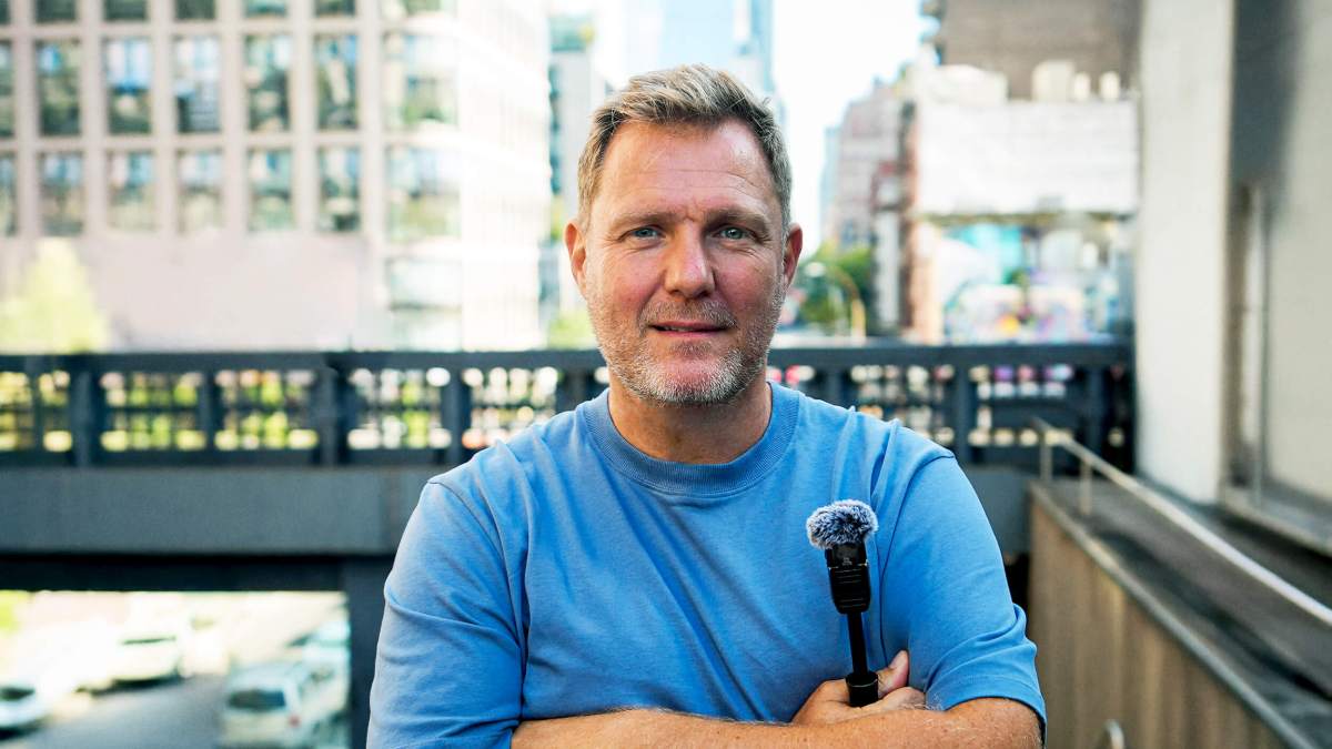 Simon Squibb, a middle-aged man in a blue shirt, stands outdoors in an urban setting, holding a small microphone with his arms crossed.
