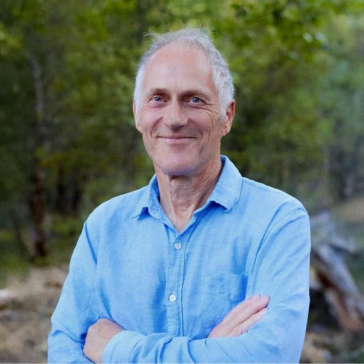 Tim O'Reilly, an older man with short gray hair wearing a light blue button-up shirt, stands outdoors with arms crossed, surrounded by green trees.