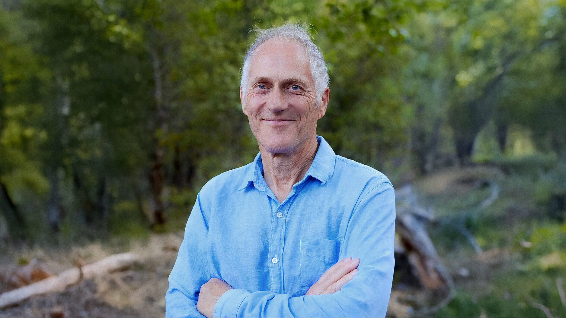 Tim O'Reilly, an older man with short gray hair wearing a light blue button-up shirt, stands outdoors with arms crossed, surrounded by green trees.